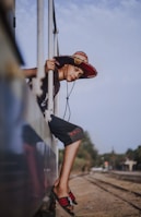 A person wearing a large, colorful straw hat leans out of a train window. The individual is wearing casual clothes and sandals, capturing a relaxed and adventurous vibe as they look into the distance. The background shows train tracks and a slightly blurred landscape with trees under a cloudy sky.