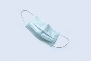 A single disposable light blue surgical face mask is placed against a plain white background. The mask features ear loops and multiple layers of fabric, typical of medical masks.