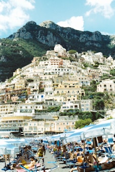 A picturesque coastal town with numerous pastel-colored buildings stacked on a steep hillside. In the foreground, a beach scene with people relaxing on sunbeds under white umbrellas. The mountainous backdrop adds to the scenic beauty, with lush greenery and rocky peaks.