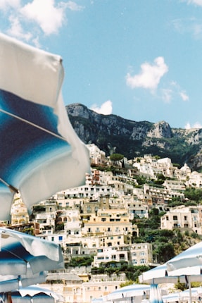 A picturesque coastal town with colorful buildings stacked on a hillside. In the foreground, there are several beach umbrellas, mostly in shades of blue and white. The ocean isn't visible, but the sky is clear and blue with a few fluffy clouds. The overall scenery is vibrant and Mediterranean.
