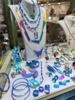 A display of various colorful jewelry pieces, including necklaces, bracelets, rings, and earrings, featuring shades of blue, purple, and green. The items are elegantly arranged on a mannequin and a showcase counter, highlighting the intricate designs and vibrant hues.