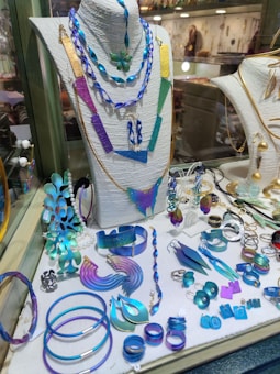 A display of various colorful jewelry pieces, including necklaces, bracelets, rings, and earrings, featuring shades of blue, purple, and green. The items are elegantly arranged on a mannequin and a showcase counter, highlighting the intricate designs and vibrant hues.