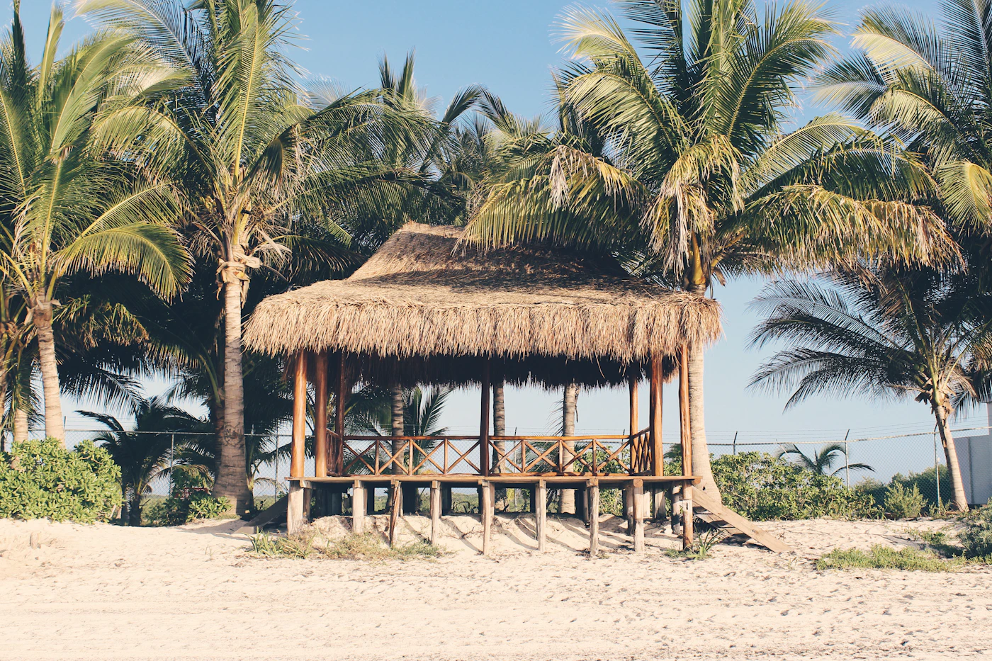 A standalone palapa beach venue — a thatched-roof open structure on the sand among coconut palms, the Riviera Maya architectural vocabulary of natural materials and open-air design