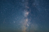 A star-filled night sky with a visible Milky Way galaxy stretching across the horizon. Countless stars are scattered throughout the dark expanse with a shooting star visible in the frame.