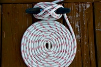 A coiled white rope with red patterns is neatly arranged on a wooden surface. The rope is looped around a black cleat, creating a symmetrical and organized appearance. The wooden background has a rustic texture with visible grains and signs of wear.