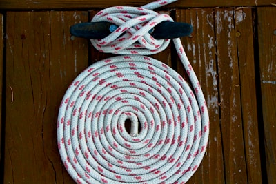 A coiled white rope with red patterns is neatly arranged on a wooden surface. The rope is looped around a black cleat, creating a symmetrical and organized appearance. The wooden background has a rustic texture with visible grains and signs of wear.