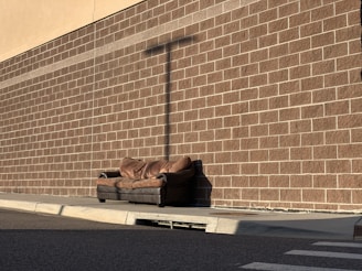 A worn brown sofa is placed on the sidewalk against a brick wall. The bricks are aligned in a grid pattern, casting a noticeable shadow from a utility pole. The area is empty, and the lighting suggests it's either early morning or late afternoon.