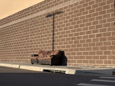 A worn brown sofa is placed on the sidewalk against a brick wall. The bricks are aligned in a grid pattern, casting a noticeable shadow from a utility pole. The area is empty, and the lighting suggests it's either early morning or late afternoon.
