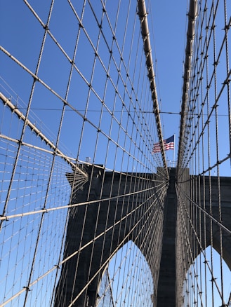 Symmetrical cables stretch across a large suspension bridge with a clear blue sky in the background. An American flag flutters at the top of one of the bridge's towers.