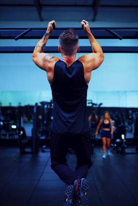 A man is performing a pull-up exercise on a bar with strong, muscular arms. He is wearing a sleeveless shirt and athletic pants. In the background, a gym setting is visible with fitness equipment and another person walking.