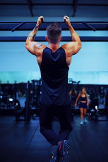 A man is performing a pull-up exercise on a bar with strong, muscular arms. He is wearing a sleeveless shirt and athletic pants. In the background, a gym setting is visible with fitness equipment and another person walking.