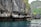 Massive limestone cliffs rise dramatically from the turquoise water, with jagged formations and patches of lush green vegetation clinging to the rock face. The rugged texture of the rocks creates a stark contrast with the calm, reflective water below.