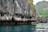 Massive limestone cliffs rise dramatically from the turquoise water, with jagged formations and patches of lush green vegetation clinging to the rock face. The rugged texture of the rocks creates a stark contrast with the calm, reflective water below.