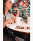 A woman is smiling and sitting at a round table with white mugs. There is a colorful brick wall in the background with red, blue, and green sections. Two mugs and a small potted plant are placed on the table along with scrabble tiles spelling 'LADO B'. A microphone is also visible on the table.