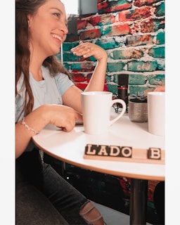 A woman is smiling and sitting at a round table with white mugs. There is a colorful brick wall in the background with red, blue, and green sections. Two mugs and a small potted plant are placed on the table along with scrabble tiles spelling 'LADO B'. A microphone is also visible on the table.