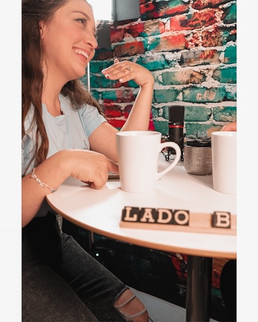 A woman is smiling and sitting at a round table with white mugs. There is a colorful brick wall in the background with red, blue, and green sections. Two mugs and a small potted plant are placed on the table along with scrabble tiles spelling 'LADO B'. A microphone is also visible on the table.