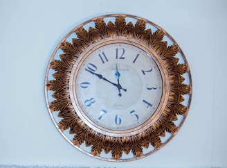 A large, round wall clock with Roman numerals and a decorative floral pattern around the face. The clock hands are ornate, styled with intricate details. The frame is embellished with a pattern resembling leaves, giving it an antique and elegant look.