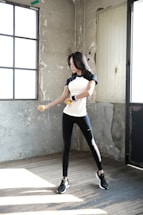 A woman is exercising with dumbbells inside a rustic room. She is wearing a black and white athletic outfit and appears focused on her workout. The room has a worn and industrial look with concrete walls and large windows that let in natural light.
