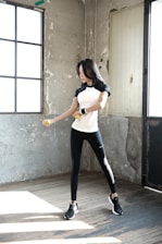 A woman is exercising with dumbbells inside a rustic room. She is wearing a black and white athletic outfit and appears focused on her workout. The room has a worn and industrial look with concrete walls and large windows that let in natural light.