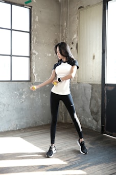 A woman is exercising with dumbbells inside a rustic room. She is wearing a black and white athletic outfit and appears focused on her workout. The room has a worn and industrial look with concrete walls and large windows that let in natural light.