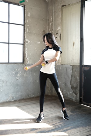 A woman is exercising with dumbbells inside a rustic room. She is wearing a black and white athletic outfit and appears focused on her workout. The room has a worn and industrial look with concrete walls and large windows that let in natural light.