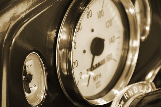 Close-up of a vintage car's speedometer and fuel gauge, featuring a classic design with a needle indicating speed in miles per hour. The dashboard elements are surrounded by metallic rings, and the overall color tone is sepia, enhancing the nostalgic feel.