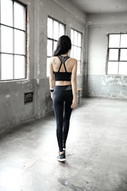 A person with long dark hair is wearing a black sports outfit, consisting of a sports bra and leggings. They are standing in a spacious, industrial-style room with tall windows that let in natural light. The walls are gray and slightly worn, adding a rustic charm to the setting.