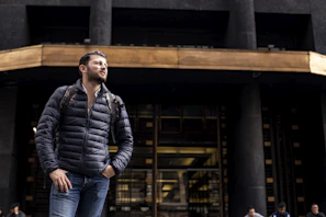 Plus size man wearing fit black jeans and a grey jacket, standing confidently in front of a modern building.