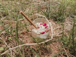 A glass with a bamboo straw, filled with a drink containing ice cubes, flowers, and seeds, rests on grass and dry straw in an outdoor setting.