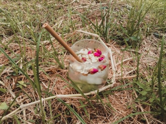 A glass with a bamboo straw, filled with a drink containing ice cubes, flowers, and seeds, rests on grass and dry straw in an outdoor setting.