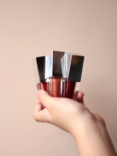 A hand holding three metallic-capped bottles, each containing a different shade of liquid lipstick or gloss. The bottles are vertically aligned, showcasing shades of red and brown. The background is a plain, soft beige color.