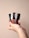 A hand holding three metallic-capped bottles, each containing a different shade of liquid lipstick or gloss. The bottles are vertically aligned, showcasing shades of red and brown. The background is a plain, soft beige color.