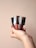 A hand holding three metallic-capped bottles, each containing a different shade of liquid lipstick or gloss. The bottles are vertically aligned, showcasing shades of red and brown. The background is a plain, soft beige color.
