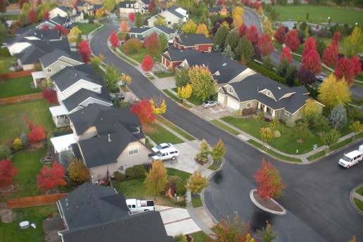 An aerial view of a suburban neighborhood featuring multiple houses with dark roofs, surrounded by streets and vibrant autumn-colored trees. The setting shows a curving layout of roads and properties, with patches of lush green lawns.
