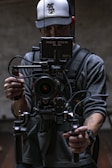 A person in a baseball cap and gray shirt is holding a professional video camera setup, which includes a RED PRO 5.0 monitor. The camera is mounted on a stabilizing rig, and various cables are connected to the equipment. The background is dimly lit, creating a moody atmosphere.