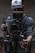 A person in a baseball cap and gray shirt is holding a professional video camera setup, which includes a RED PRO 5.0 monitor. The camera is mounted on a stabilizing rig, and various cables are connected to the equipment. The background is dimly lit, creating a moody atmosphere.