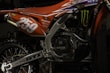 A close-up view of a motorcycle's engine and rear section painted in orange and adorned with various decals. The number '259' is prominently displayed in white. The intricate details of the metallic components and the rugged tire are visible, along with brand logos such as Monster Energy.