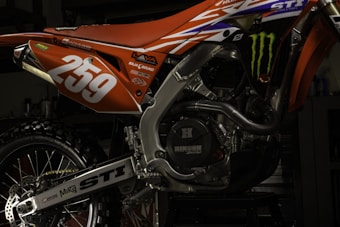 A close-up view of a motorcycle's engine and rear section painted in orange and adorned with various decals. The number '259' is prominently displayed in white. The intricate details of the metallic components and the rugged tire are visible, along with brand logos such as Monster Energy.