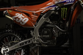 A close-up view of a motorcycle's engine and rear section painted in orange and adorned with various decals. The number '259' is prominently displayed in white. The intricate details of the metallic components and the rugged tire are visible, along with brand logos such as Monster Energy.