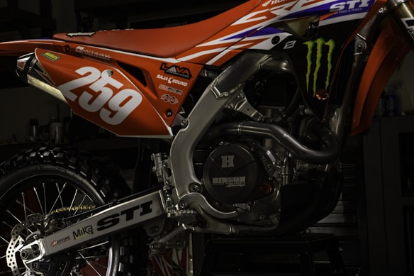 A close-up view of a motorcycle's engine and rear section painted in orange and adorned with various decals. The number '259' is prominently displayed in white. The intricate details of the metallic components and the rugged tire are visible, along with brand logos such as Monster Energy.