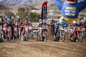 A group of motocross riders is lined up at the starting line, ready to race on a dirt track. Each rider is equipped with colorful gear and helmets, displaying their racing numbers prominently on the front of their bikes. The background features a large inflatable figure holding a sign about the weekend race, along with a mountainous desert landscape and spectators.