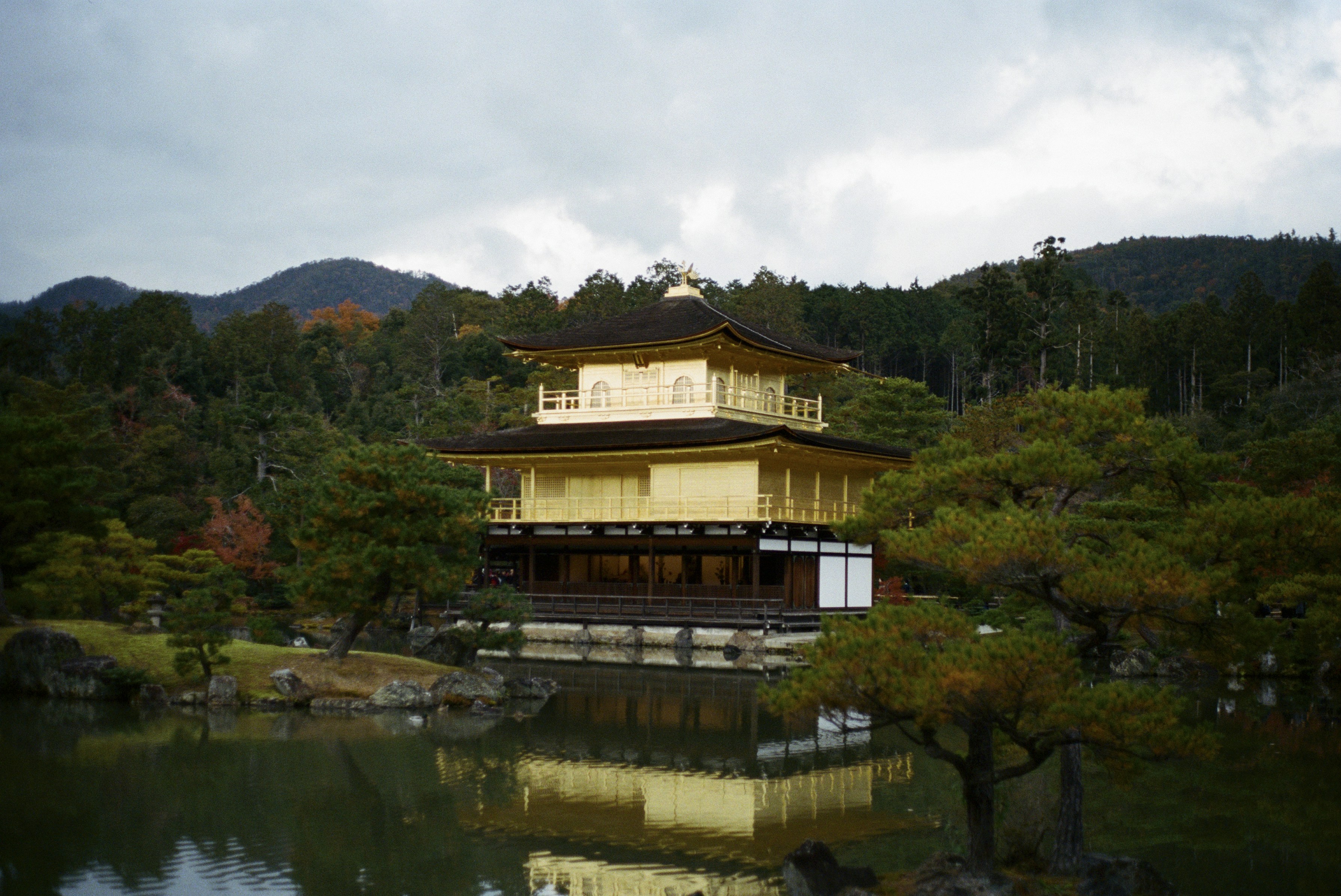 Gold leaf temple sits on a tranquil pond, its mirrored facade framed by evergreen pines and distant hills.
