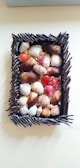 A wooden frame filled with an assortment of seashells, varying in color and size. The frame is constructed from dark, twig-like pieces forming a rectangular border. Inside the frame, the seashells are arranged closely together, displaying hues of orange, red, white, and brown.