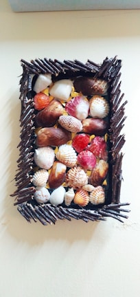 A wooden frame filled with an assortment of seashells, varying in color and size. The frame is constructed from dark, twig-like pieces forming a rectangular border. Inside the frame, the seashells are arranged closely together, displaying hues of orange, red, white, and brown.