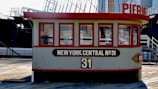 A vintage-looking building structure with multiple windows and a sign reading 'New York Central No.31'. This structure appears to be situated on a wooden deck. There's a red and cream color scheme in the building, with industrial and maritime elements like ship masts in the background.