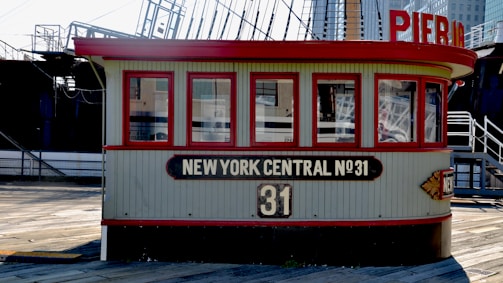 A vintage-looking building structure with multiple windows and a sign reading 'New York Central No.31'. This structure appears to be situated on a wooden deck. There's a red and cream color scheme in the building, with industrial and maritime elements like ship masts in the background.