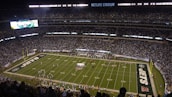 A packed football stadium with thousands of fans in attendance, centered around a brightly lit field. The field displays football markings and logos, indicating it's a professional game setup. An electronic scoreboard is visible, and the audience is engaged with the game.
