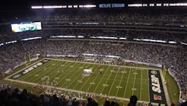 A packed football stadium with thousands of fans in attendance, centered around a brightly lit field. The field displays football markings and logos, indicating it's a professional game setup. An electronic scoreboard is visible, and the audience is engaged with the game.