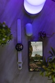 A wooden surface features a smart watch with a white band, a smartphone displaying a picturesque wallpaper of cliffs and ruins, a pair of wireless earbuds with a Samsung logo on the case, and a soft glowing light emitting a purple hue. Two small potted plants frame the scene, adding a touch of greenery.