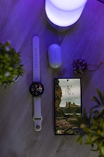 A wooden surface features a smart watch with a white band, a smartphone displaying a picturesque wallpaper of cliffs and ruins, a pair of wireless earbuds with a Samsung logo on the case, and a soft glowing light emitting a purple hue. Two small potted plants frame the scene, adding a touch of greenery.